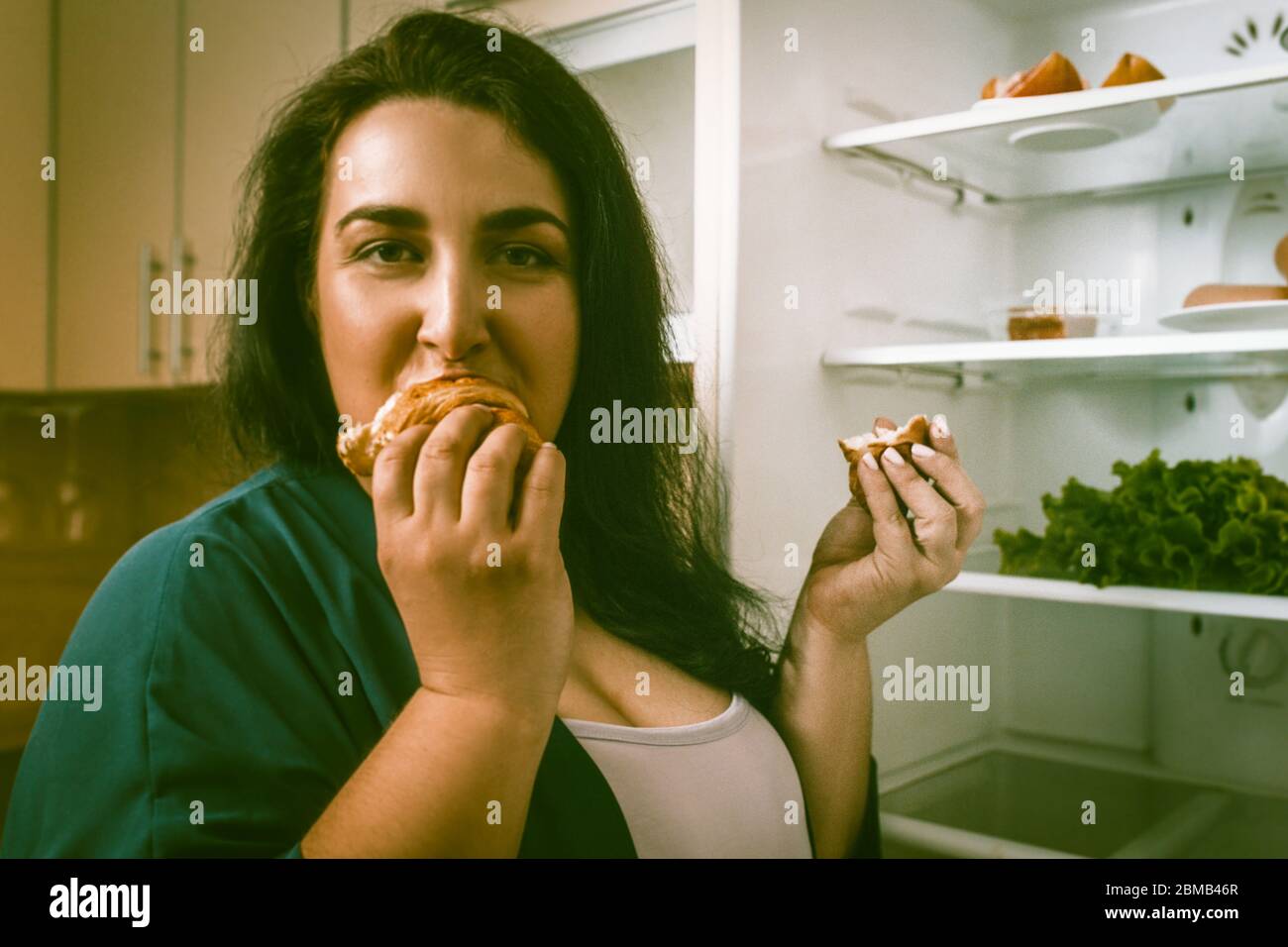 La donna in sovrappeso mangia cibo malsano, la donna affamata caucasica mangia o divora un croissant guardando la macchina fotografica Foto Stock