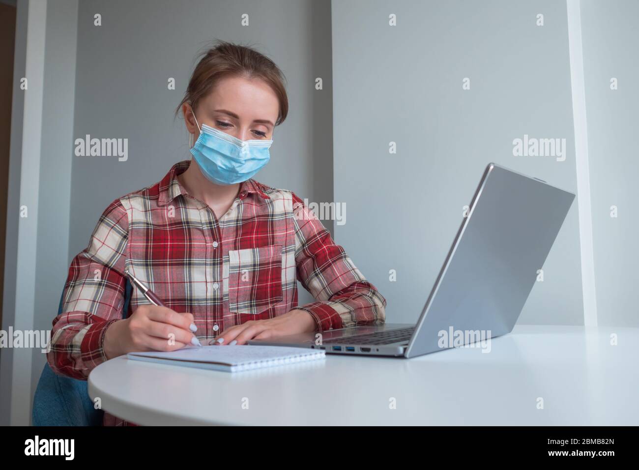 Donna in maschera medica utilizzando il computer portatile, guardando webinar, lezioni, prendere appunti in camera grigia a casa. Istruzione, freelance, internet, quarantena Foto Stock