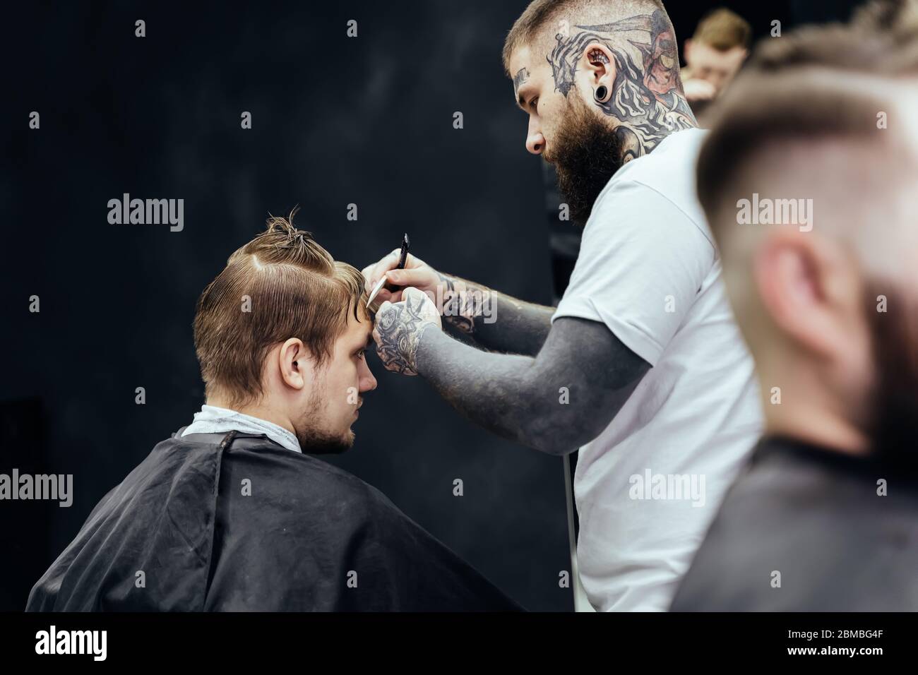 Barbiere professionale tatuato con rasoio a gola di taglio. Un maschio attraente sta ottenendo un taglio moderno di capelli nel barbiere. Primo piano. Foto Stock