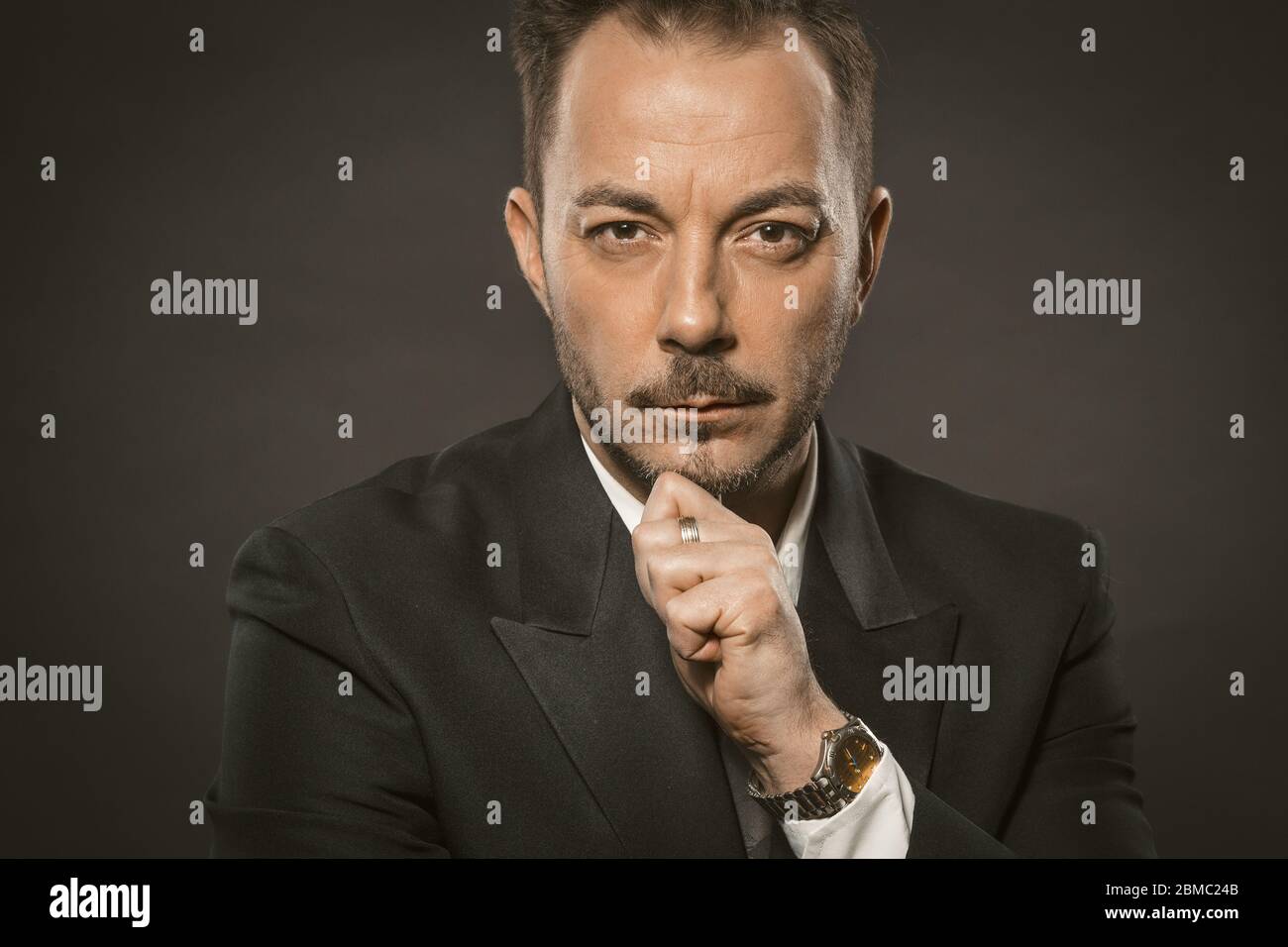 Un uomo d'affari serio tocca il mento su sfondo grigio. Ritratto di un uomo di successo fiducia in elegante formalwear guardando la fotocamera. Foto Stock