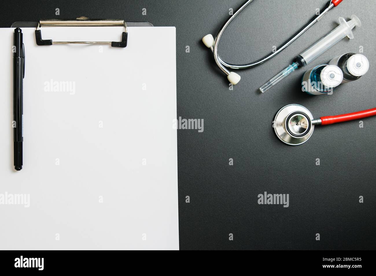 Giornata del medico e concetto di Covid-19. Lavagna a clip con fiala medica blu, iniezione e stetoscopio su tavolo scuro Foto Stock