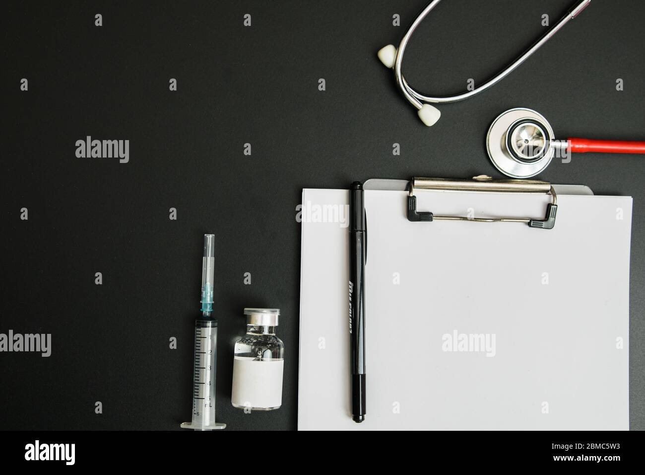 Giornata del medico e concetto di Covid-19. Lavagna a clip con fiala medica bianca, iniezione e stetoscopio su tavolo scuro Foto Stock