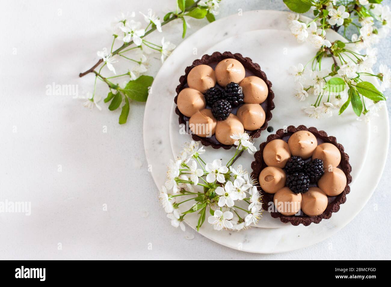 Crostata di cioccolato con ripieno di caramello salato e bacche nere fresche. Fiori di ciliegio su sfondo. Spazio di copia. Foto Stock