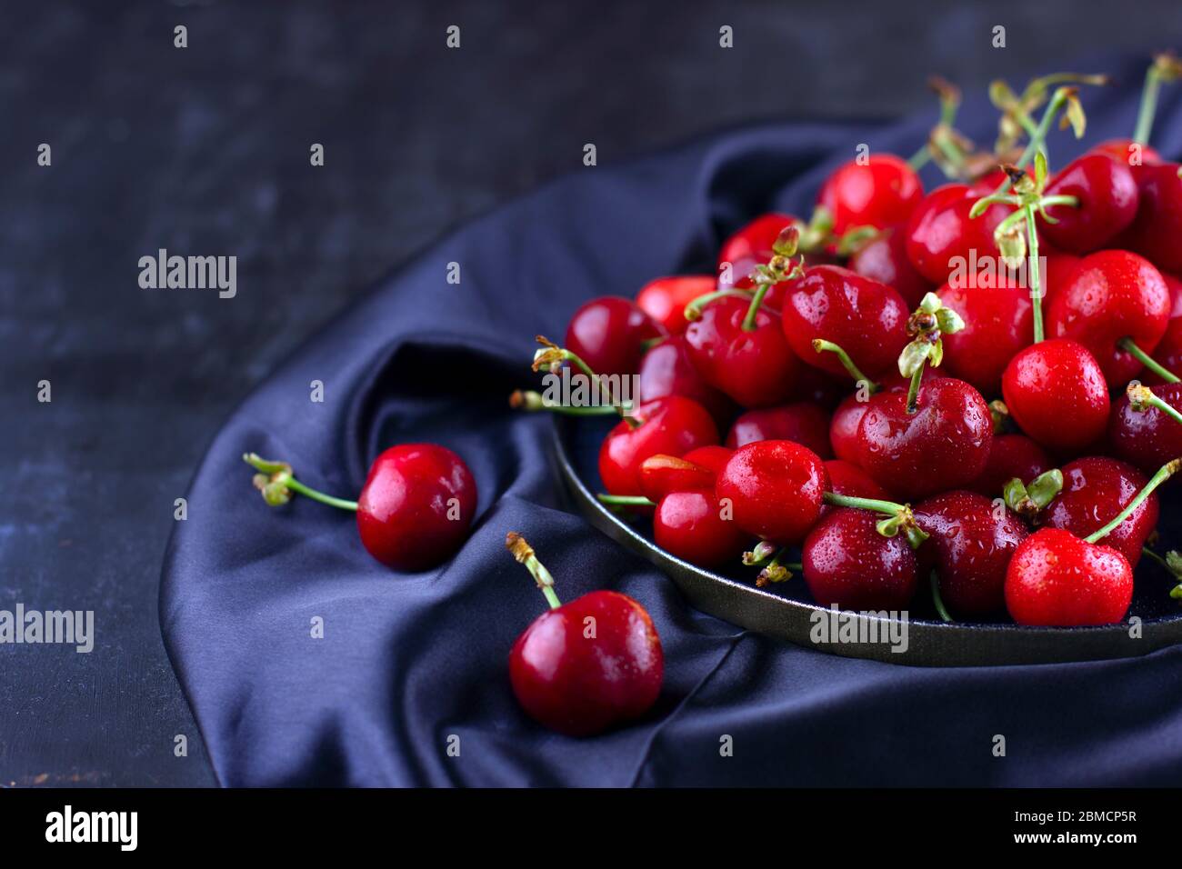 Ciliegio fresco su tessuto di seta scuro. Bacche dolci mature in gocce d'acqua Foto Stock