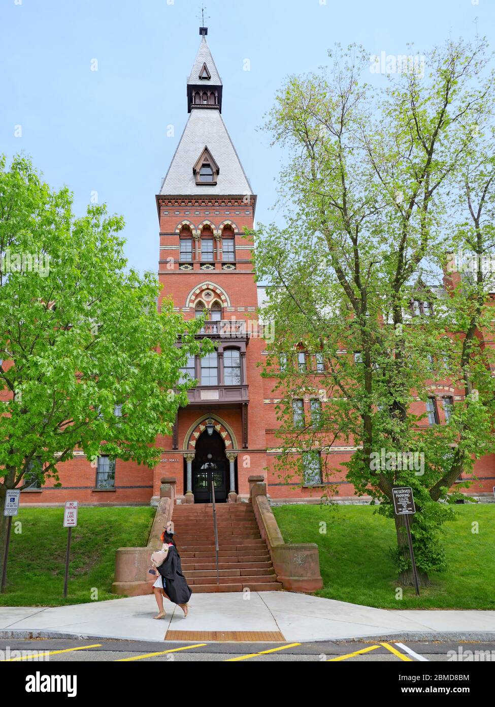 ITHACA, NY - 2019 MAGGIO: Una studentessa laureata che indossa un cappello e una passeggio passa davanti ad un edificio in stile romanico all'Università di Cornell. Foto Stock