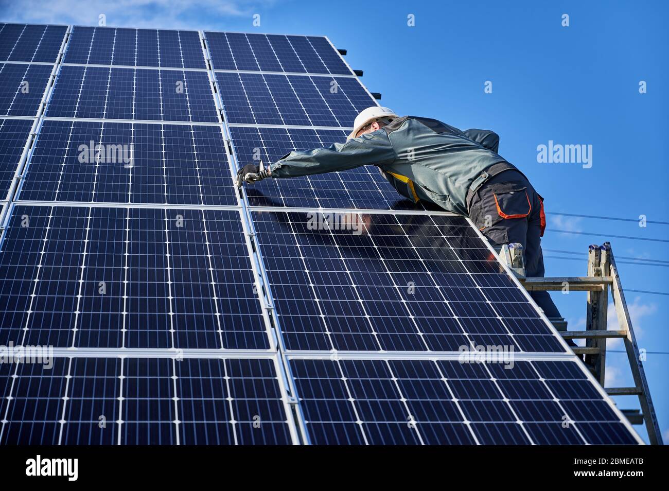 Elettricista professionista che si trova sulla scala e che installa un sistema di pannelli solari fotovoltaici. Uomo tecnico in casco di sicurezza sotto cielo blu. Concetto di energia alternativa e di potenza Foto Stock