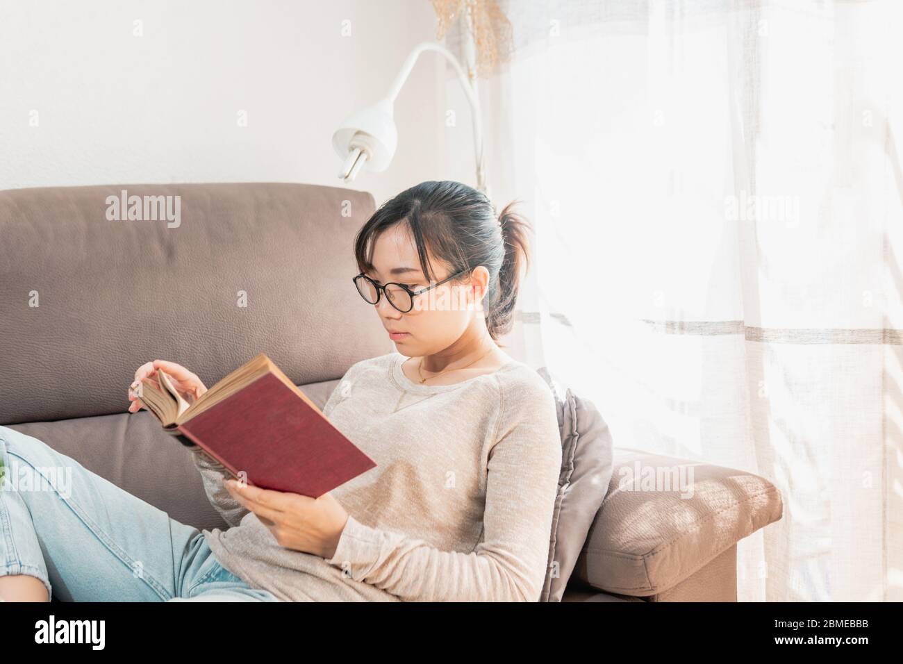 Donna asiatica che legge un libro a casa. Ragazza che si rilassa sul divano godendo un vecchio libro all'interno durante il blocco del coronavirus. Donna ha messo a fuoco la lettura. Foto Stock