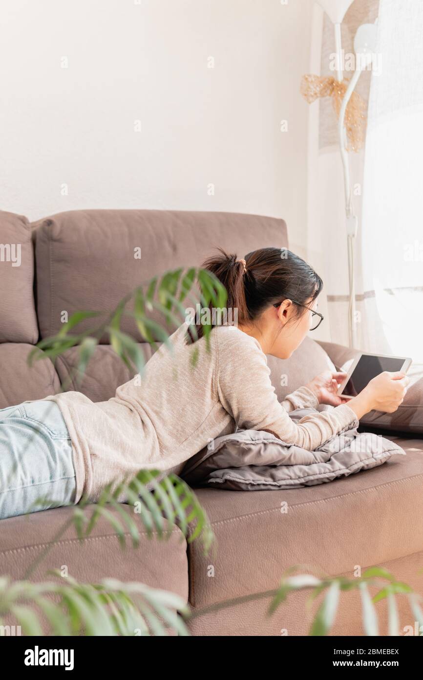 Giovane madre asiatica che si diverta da sola sdraiata sul divano guardando i video sul tablet. Ragazza che si gode a casa sul divano guardando i film. Foto Stock