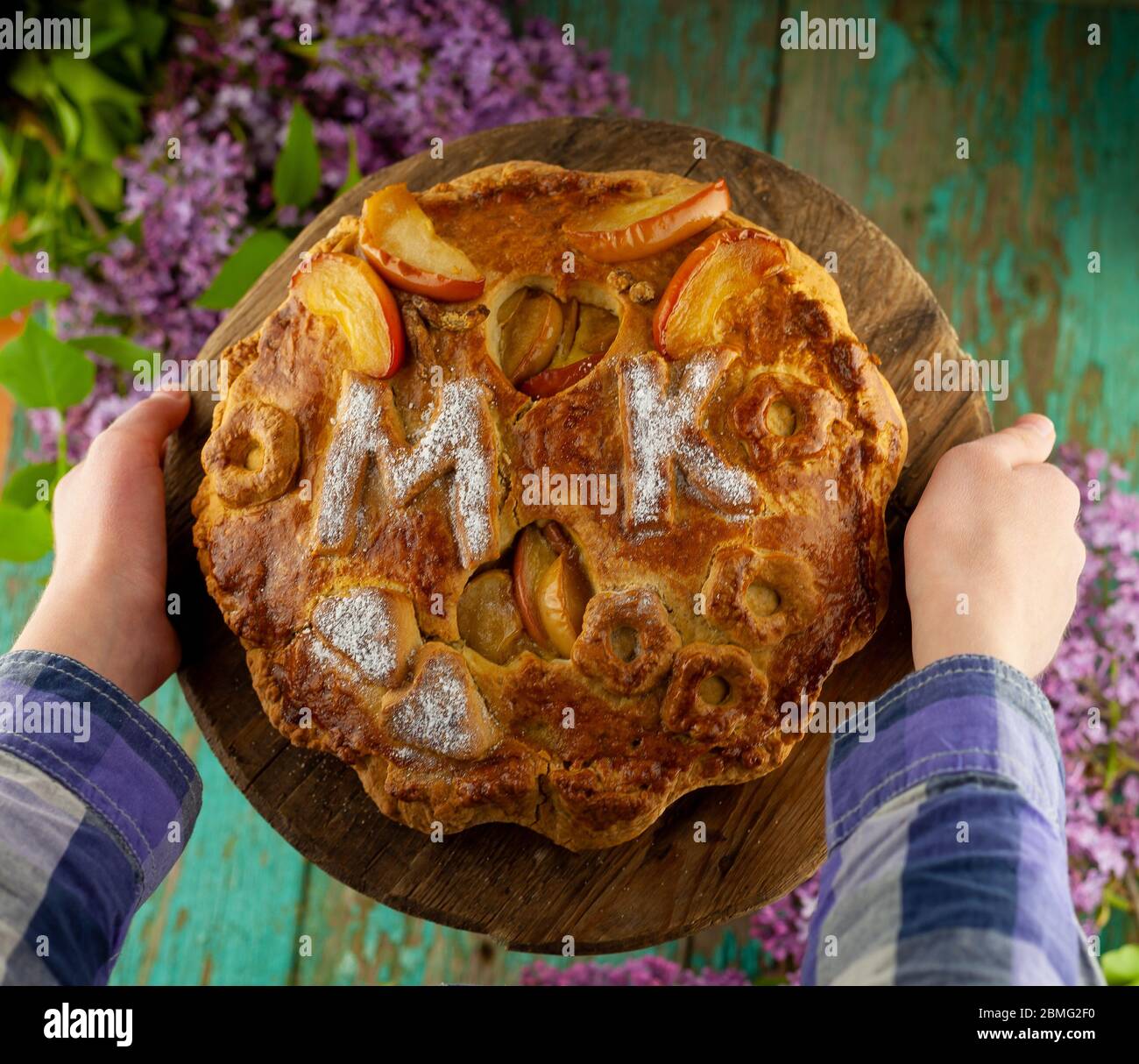 Tenere una torta rotonda fatta in casa nelle mani. Mettere sul tavolo. Un bouquet di lilacs. Le lettere MK . Semplice pasticceria fatta con pasta frolla e. Foto Stock