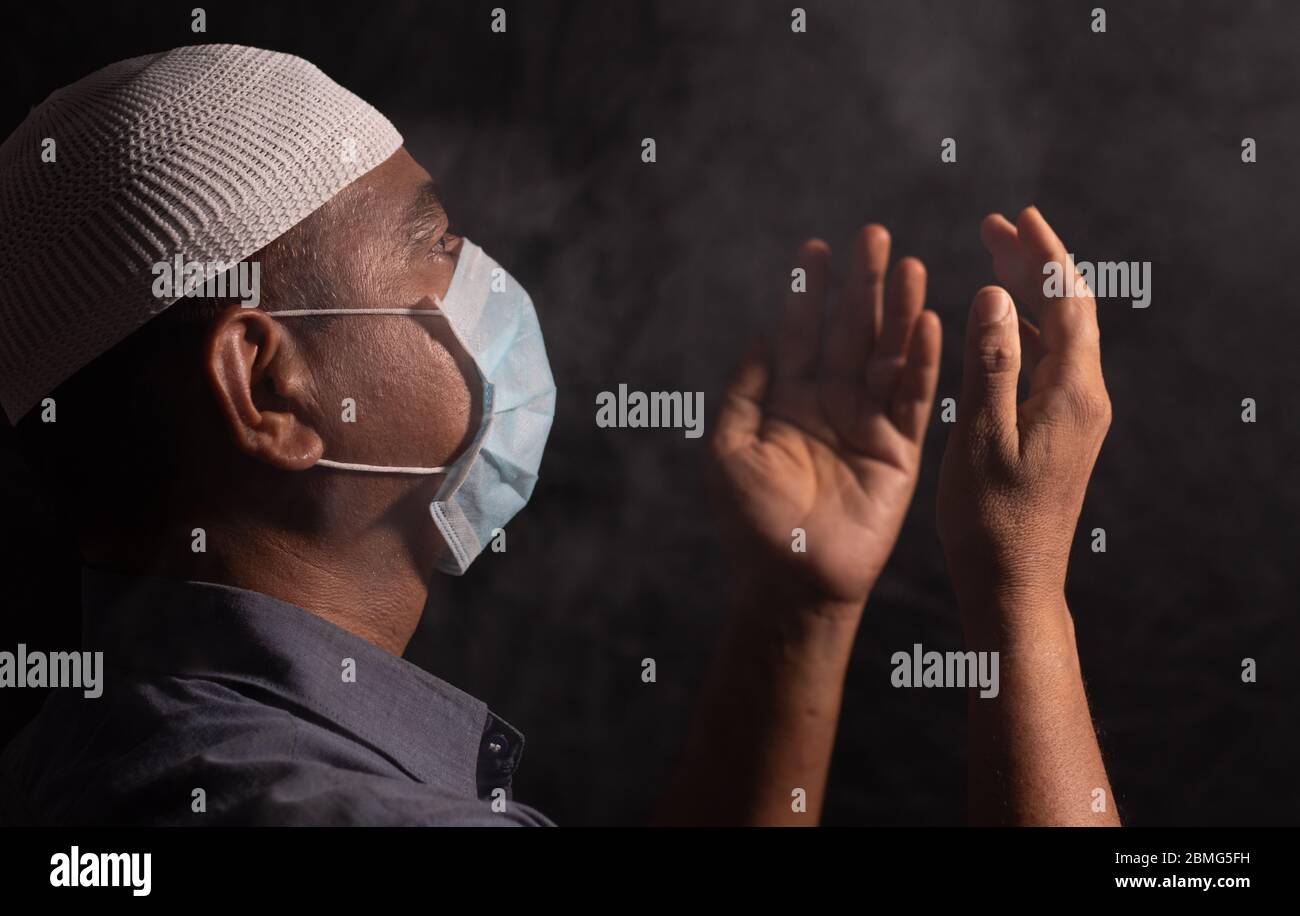 Uomo musulmano con maschera medica e cappuccio che prega a dio in stanza oscura per proteggere o salvare dal covid-19 o crisi di coronavirus - spiritualità e resa Foto Stock