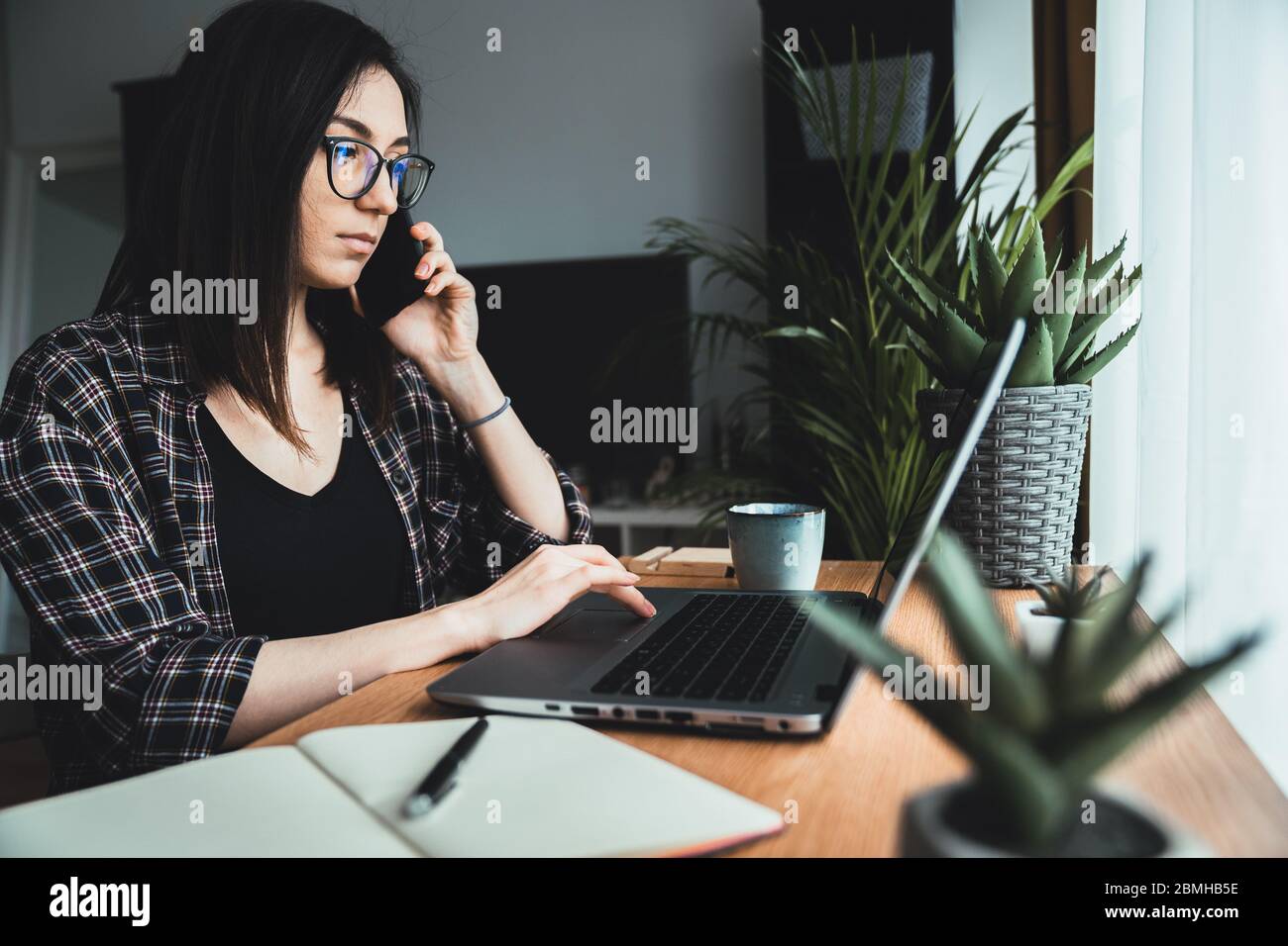 Giovane donna che parla al telefono mentre lavora al laptop. Donna d'affari in conversazione al telefono in ufficio. Concetto di lavoro da casa Foto Stock