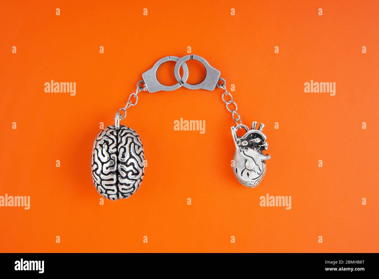 Disposizione piatta di copie anatomiche miniaturizzate del cervello e del cuore umano collegate a manette isolate su sfondo arancione. Sentimenti e connessione mentale Foto Stock