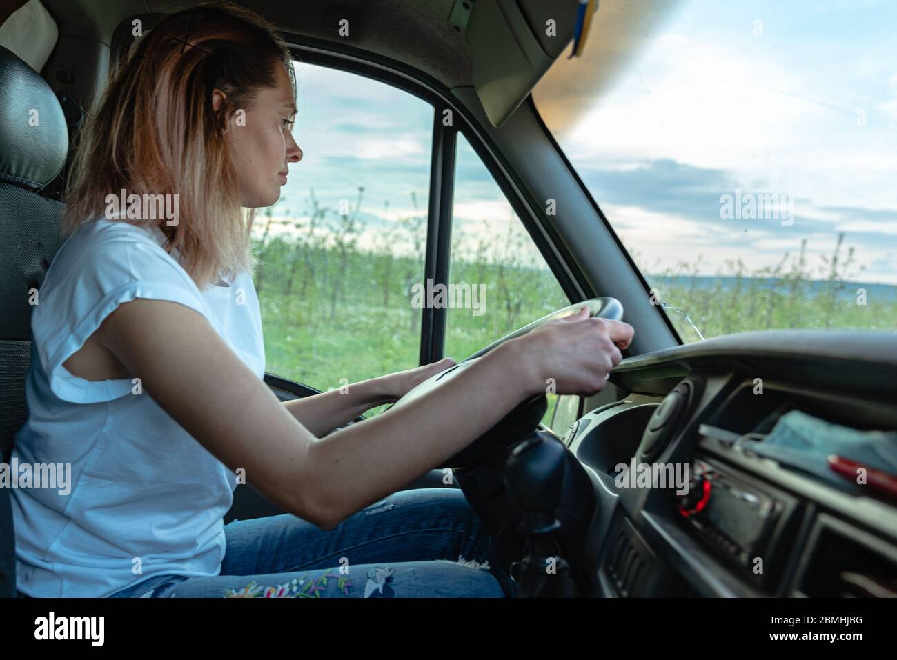 Giovane donna stanca in maglietta bianca che guida un'auto tra giardini fioriti Foto Stock