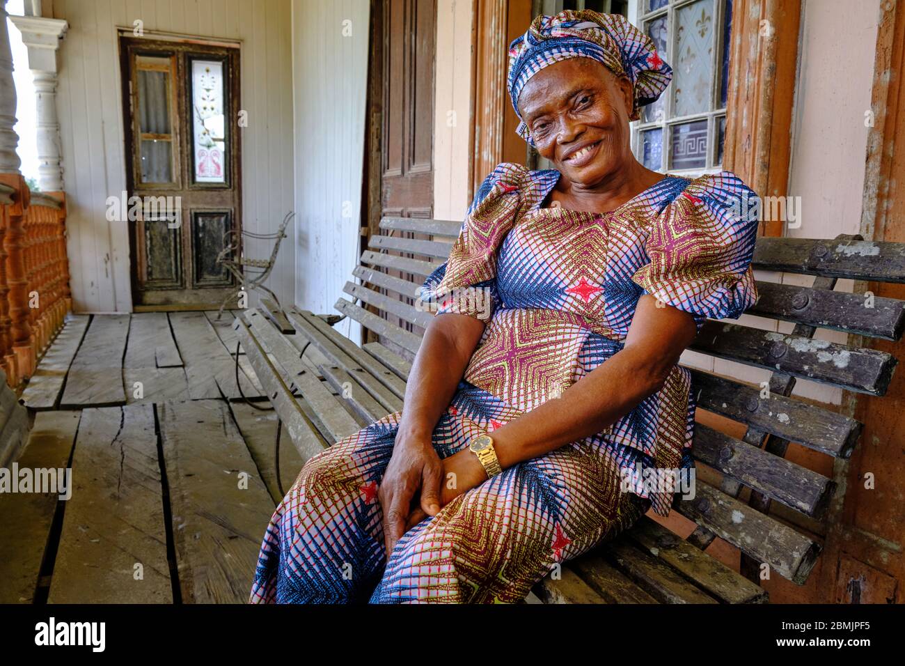 Ritratto di una donna di mezza età sorridente vestita con un colorato abito etnico seduto alle porte della sua casa coloniale Creektown. Foto Stock