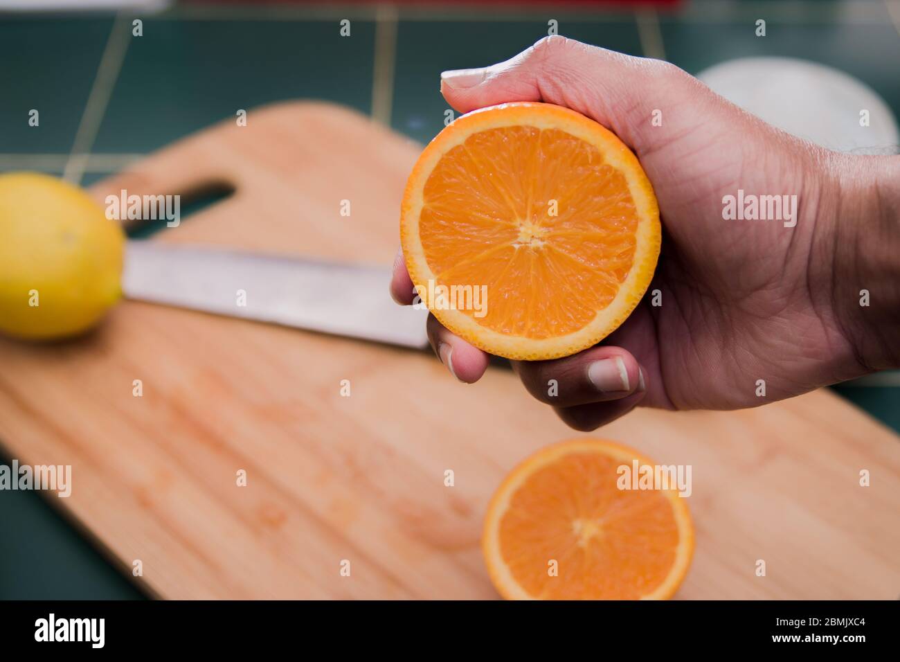 Arancio luminoso, appena tagliato, arancione che sta per essere schiacciato con una mano dell'uomo. Tagliere di legno, coltello e un limone sullo sfondo. Foto Stock