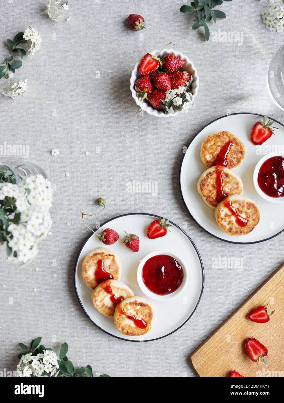 Frittelle di formaggio ucraino fatte in casa Syrniki con marmellata di fragole rosse e frutti vicino a fiori bianchi sulla vista da tavolo in stoffa Foto Stock