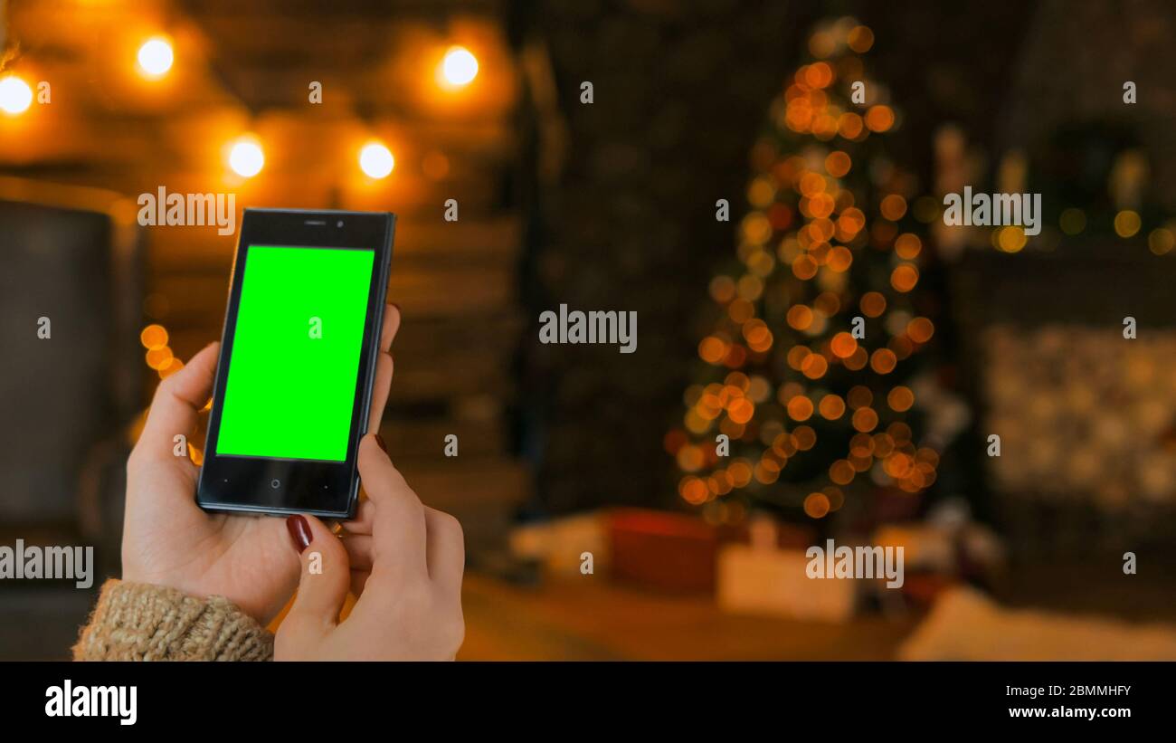 Donna che guarda uno smartphone verticale con schermo verde di notte. Natale sfondo bokeh vacanze. Mockup, modello e concetto di tecnologia Foto Stock