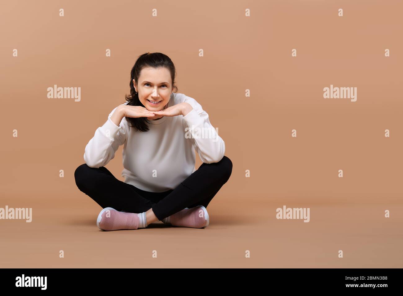Felice ragazza sorridente in bianco pullover e leggings nero seduta a gambe incrociate sul pavimento in studio Foto Stock
