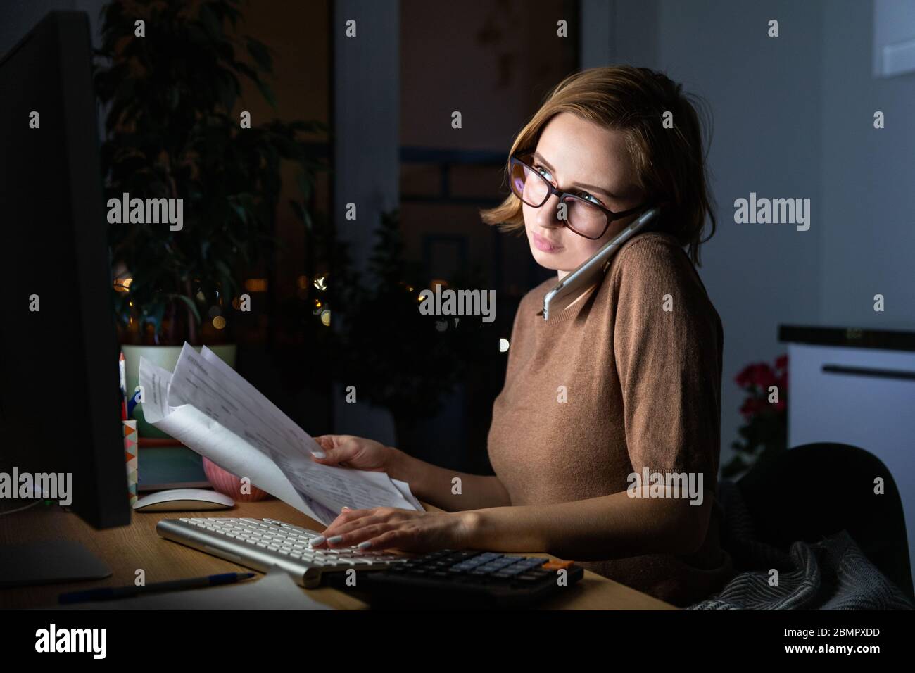 Donna in occhiali che utilizza la calcolatrice per calcolare la fattura, pianificare le spese, parlare sullo smartphone, lavorare sul PC desktop a tarda notte a casa offi Foto Stock