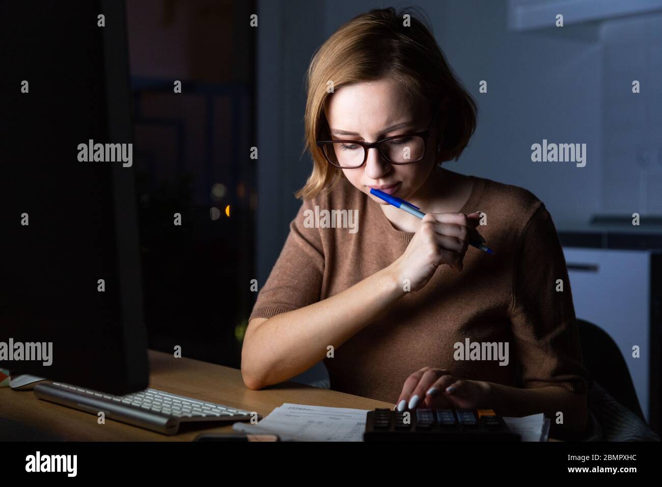 Donna in occhiali utilizzando la calcolatrice per calcolare la fattura, pianificare le spese, tenere una penna vicino al viso, lavorare su PC desktop a tarda notte a casa Foto Stock