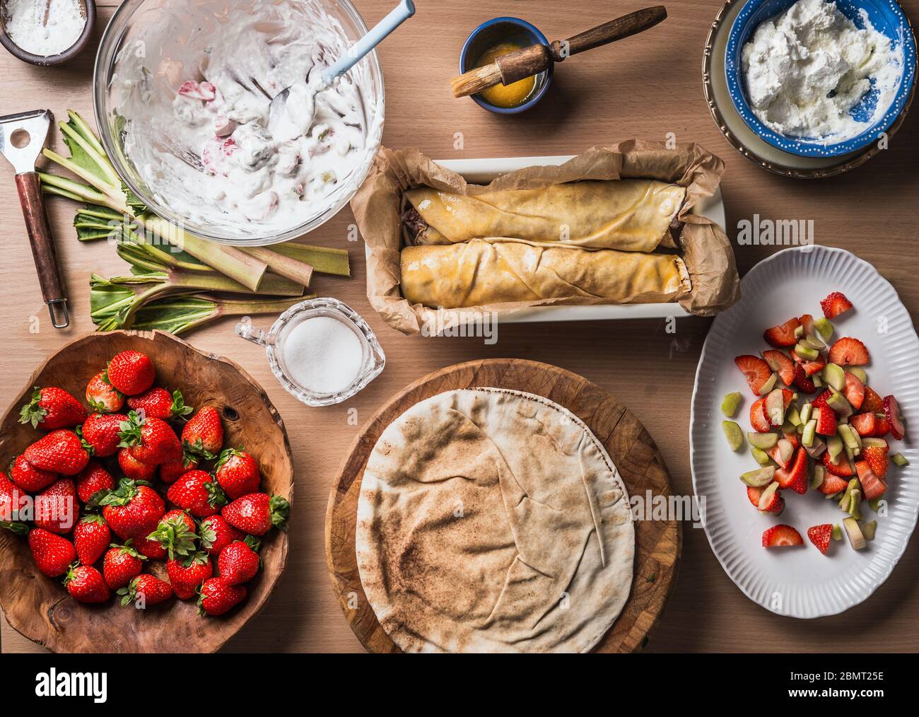 Preparazione torta di rabarbaro di fragole senza impasto, ma con pane piatto sullo sfondo del tavolo da cucina in legno con ciotole con ingredienti. Superiore Foto Stock