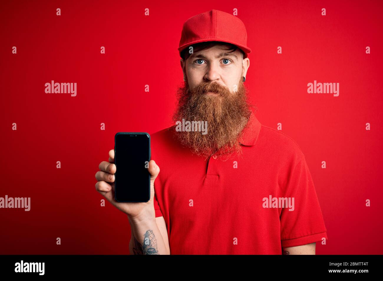 Uomo di consegna irlandese Redhead con smartphone che tiene la barba e mostra app sullo schermo con un'espressione sicura sul pensiero intelligente del viso serio Foto Stock