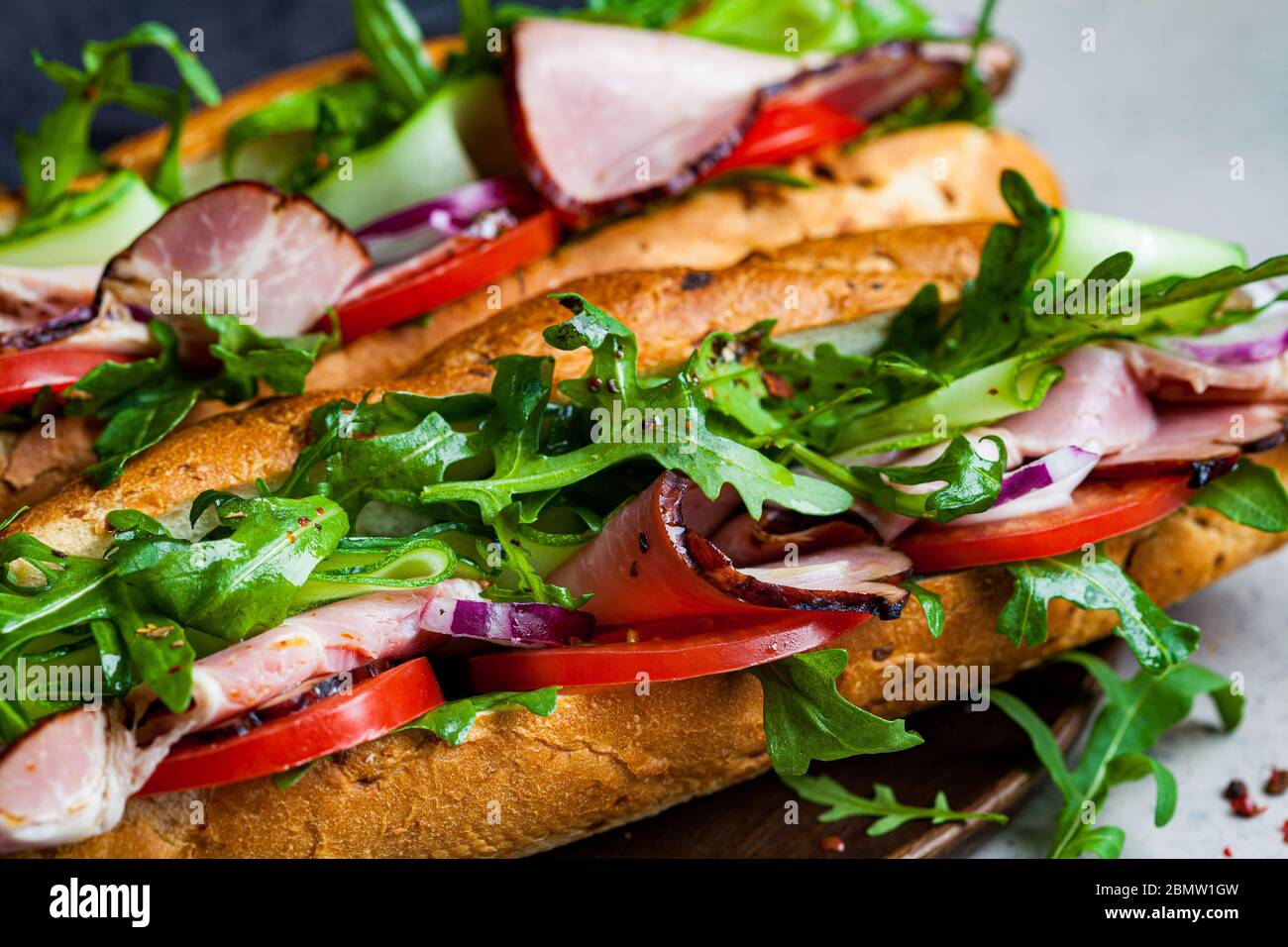 Due panini baguette freschi con carne, pomodoro, cetrioli e rucola su sfondo grigio. Foto Stock