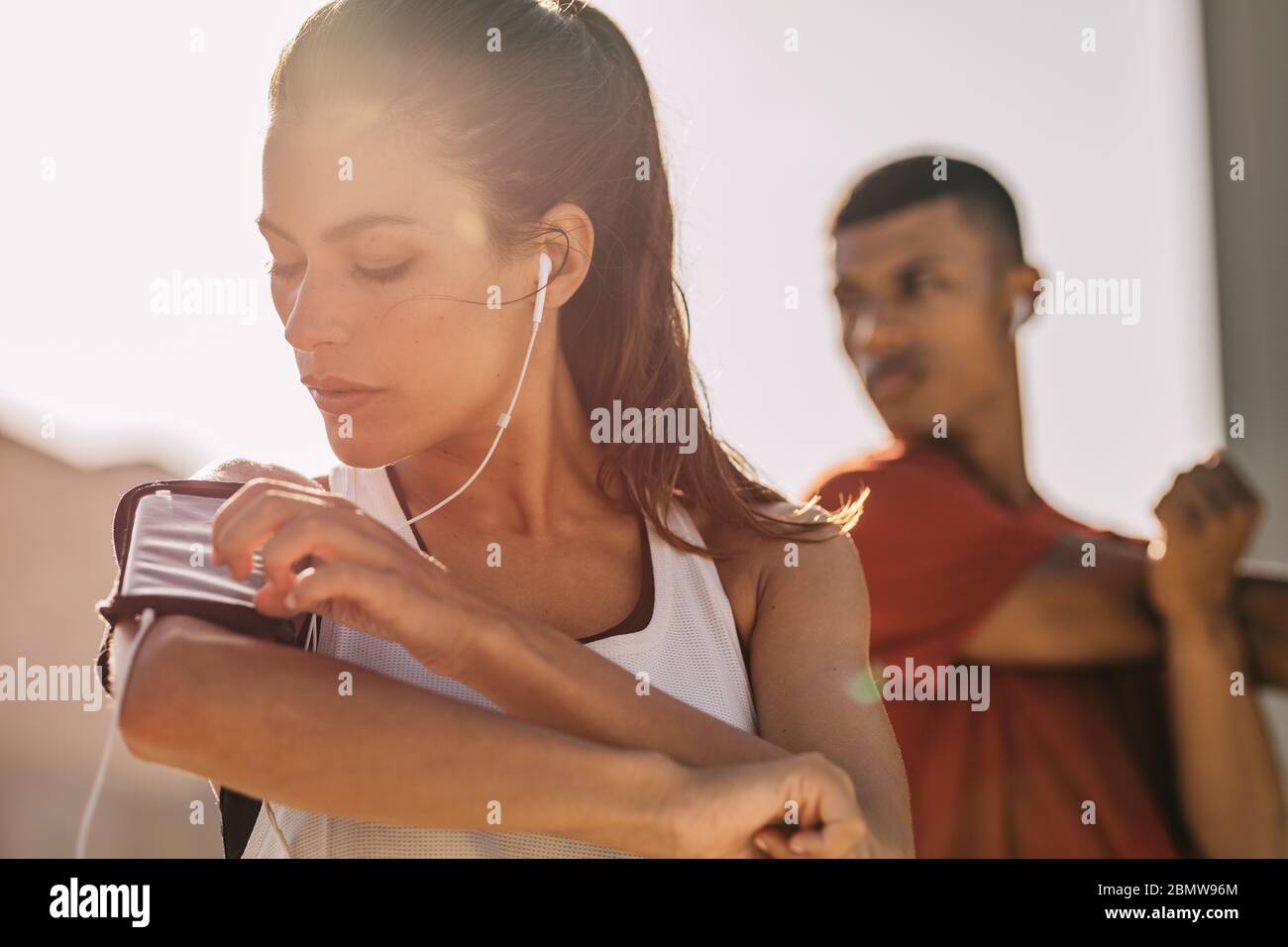 Primo piano di una donna che ascolta musica con gli auricolari dal suo smartphone mentre si esercita in città con un uomo in background. Donna che usa uno smartp Foto Stock
