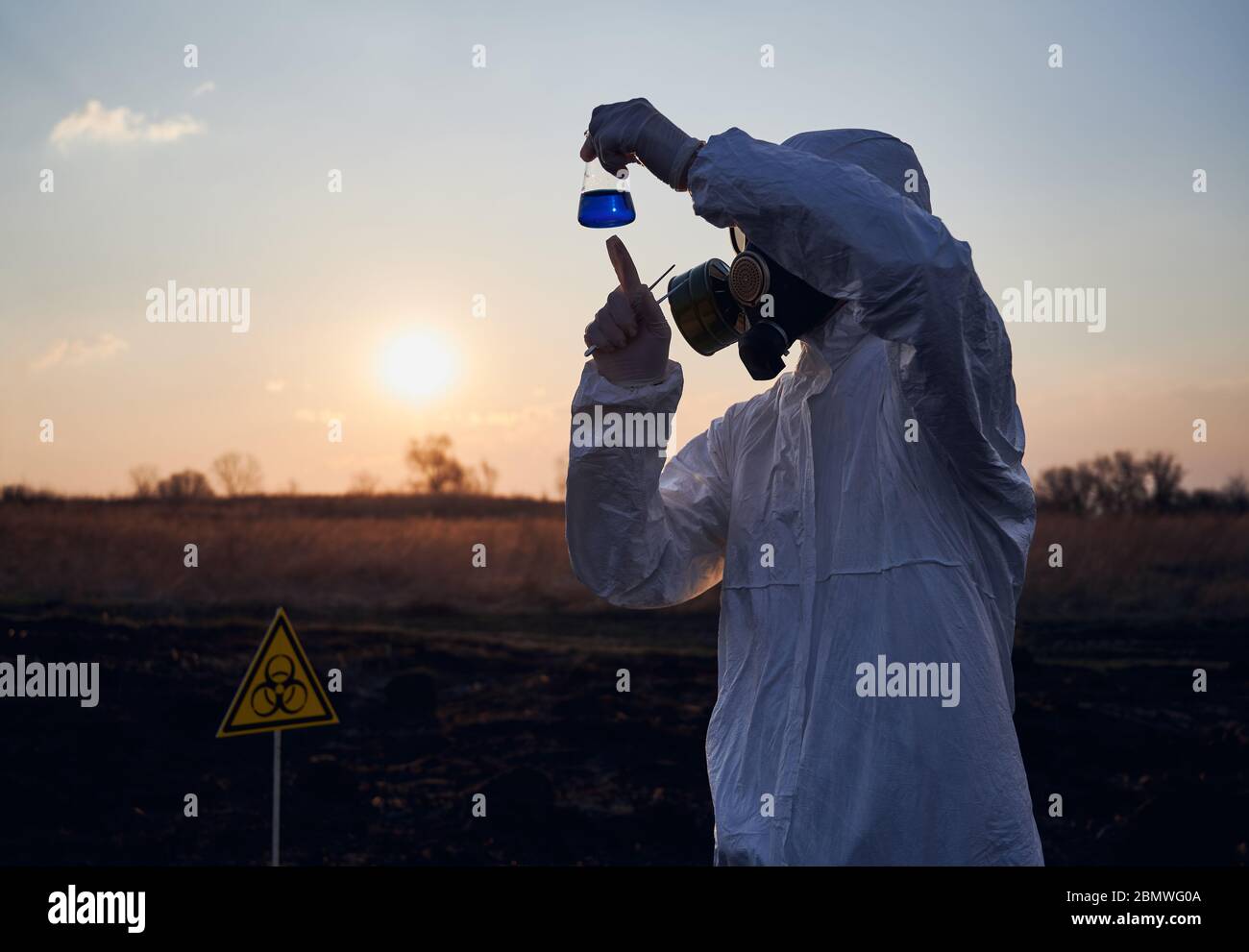 Ambientalista in tuta, maschera a gas, guanti sterili per attività di ricerca sul campo con erba bruciata, tubo di prova con liquido blu, studio di campioni di terreno su territorio bruciato con cartello a rischio biologico. Foto Stock