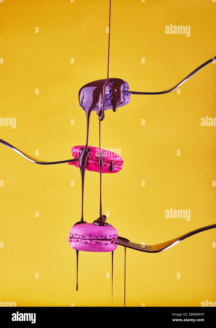 Macaron multicolore su forchette con cioccolato che scorre dall'alto su sfondo giallo Foto Stock