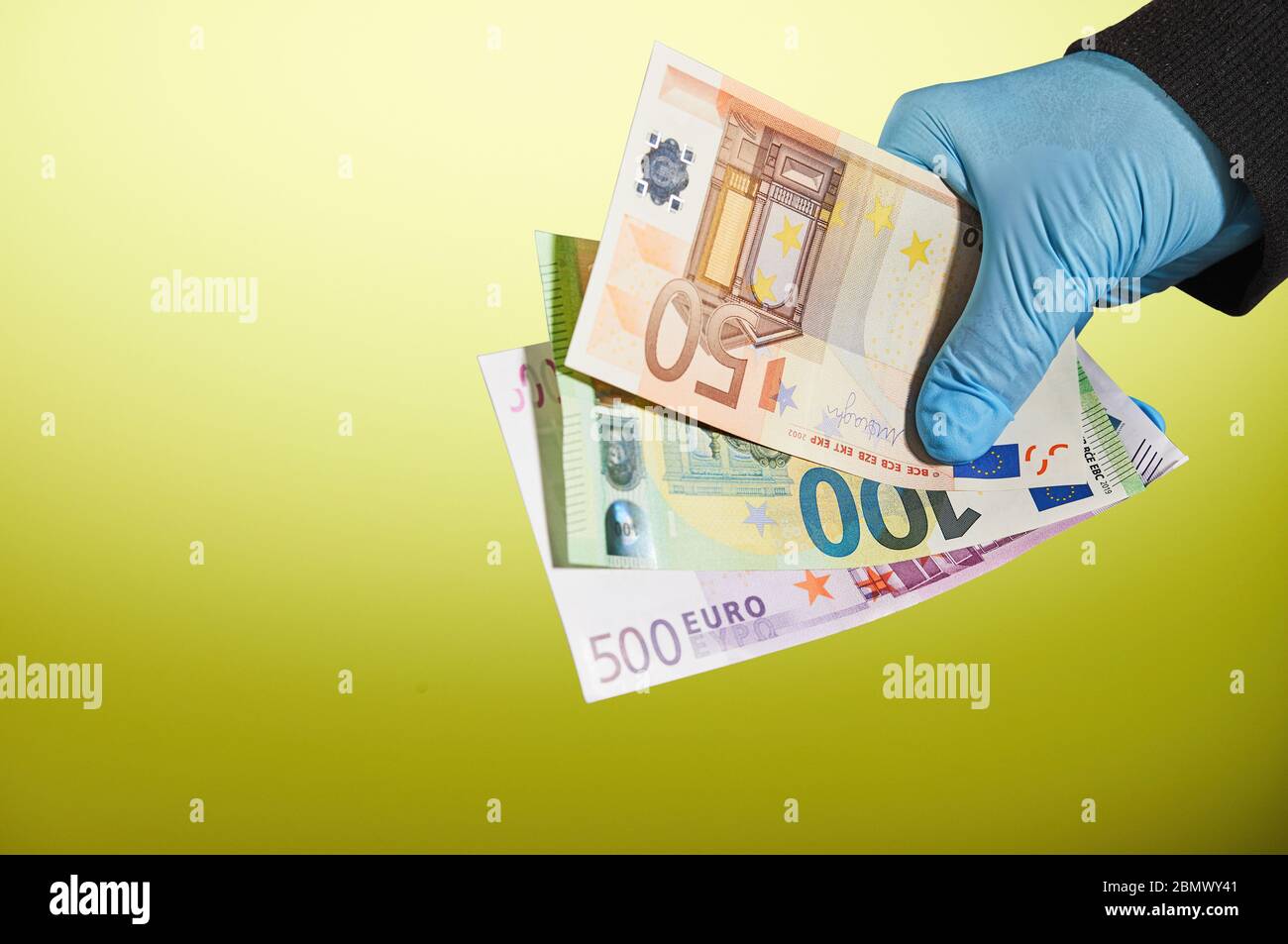 La mano di un uomo in un guanto medico blu trattiene i soldi sotto forma di banconote in euro su sfondo giallo. Protezione contro virus e batteri Foto Stock