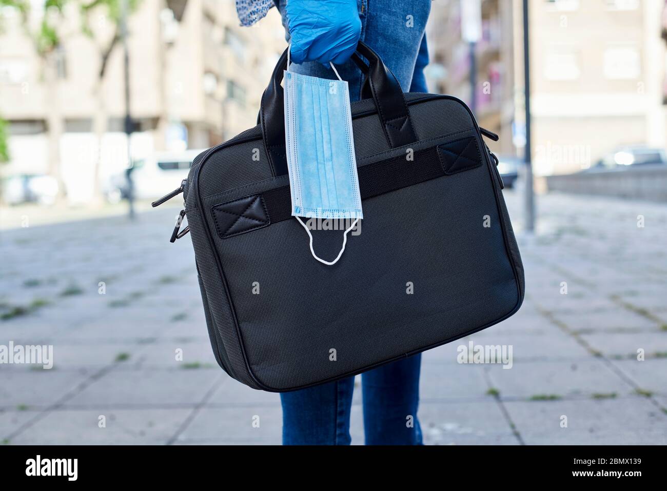 primo piano di un giovane, indossando guanti blu in lattice, portando una valigetta e una maschera chirurgica in mano sulla strada Foto Stock