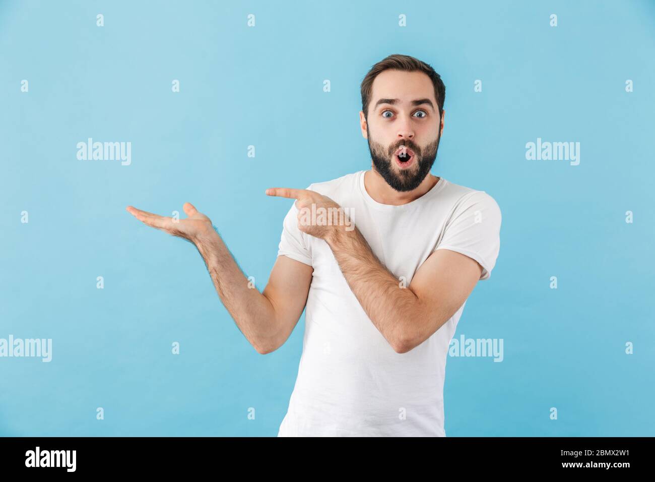 Ritratto di un giovane uomo barbuto scioccato che indossa la t-shirt in piedi isolato su sfondo blu, presentando spazio copia Foto Stock