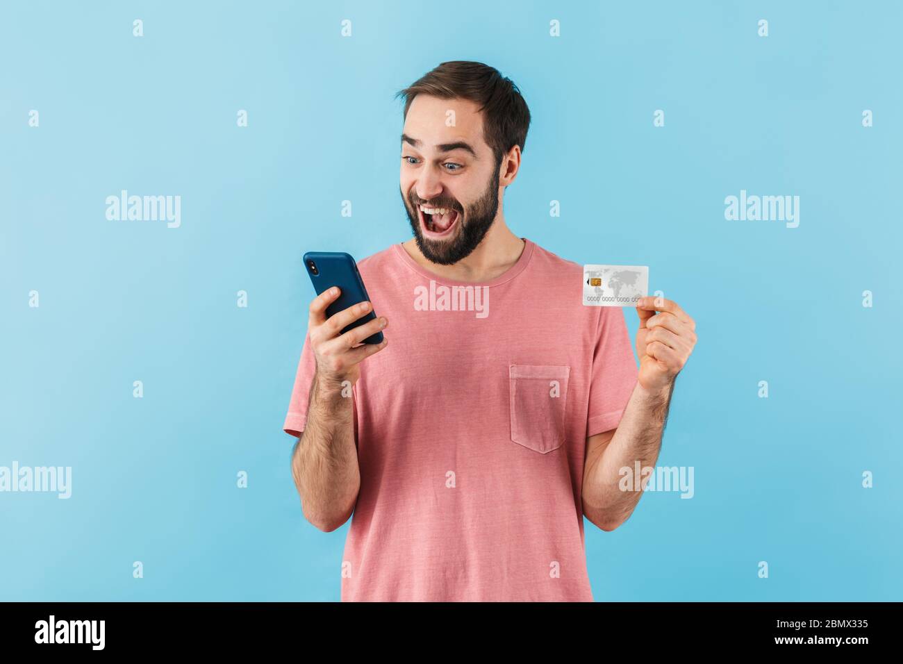 Ritratto di un giovane allegro ed eccitato uomo bearded che indossa la t-shirt in piedi isolato su sfondo blu, usando il telefono cellulare, mostrando carta di credito, negozio Foto Stock