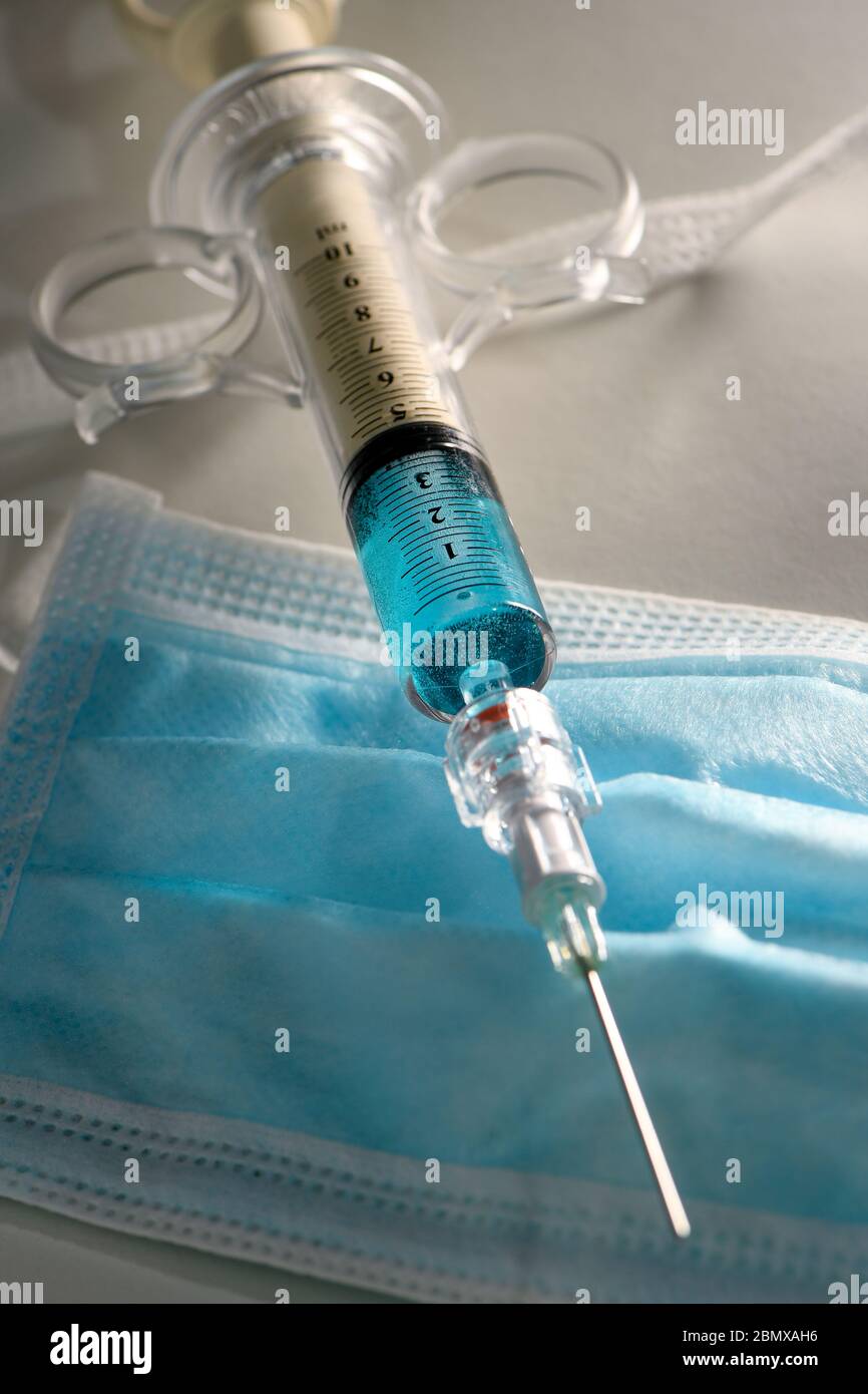 Immagine concettuale di una siringa contenente liquido blu su una maschera facciale blu DPI. Potrebbe essere usato per illustrare la pandemia di covid-19. Foto Stock