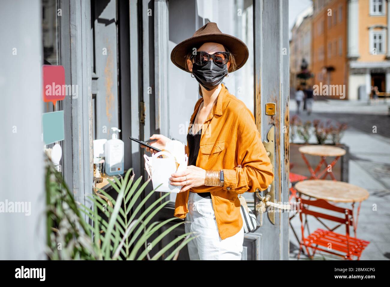 Ritratto di una giovane donna in maschera protettiva facciale in piedi con un cibo da asporto vicino all'entrata del caffè all'aperto. Concetto di nuove regole sociali per le imprese dopo la pandemia del coronavirus Foto Stock