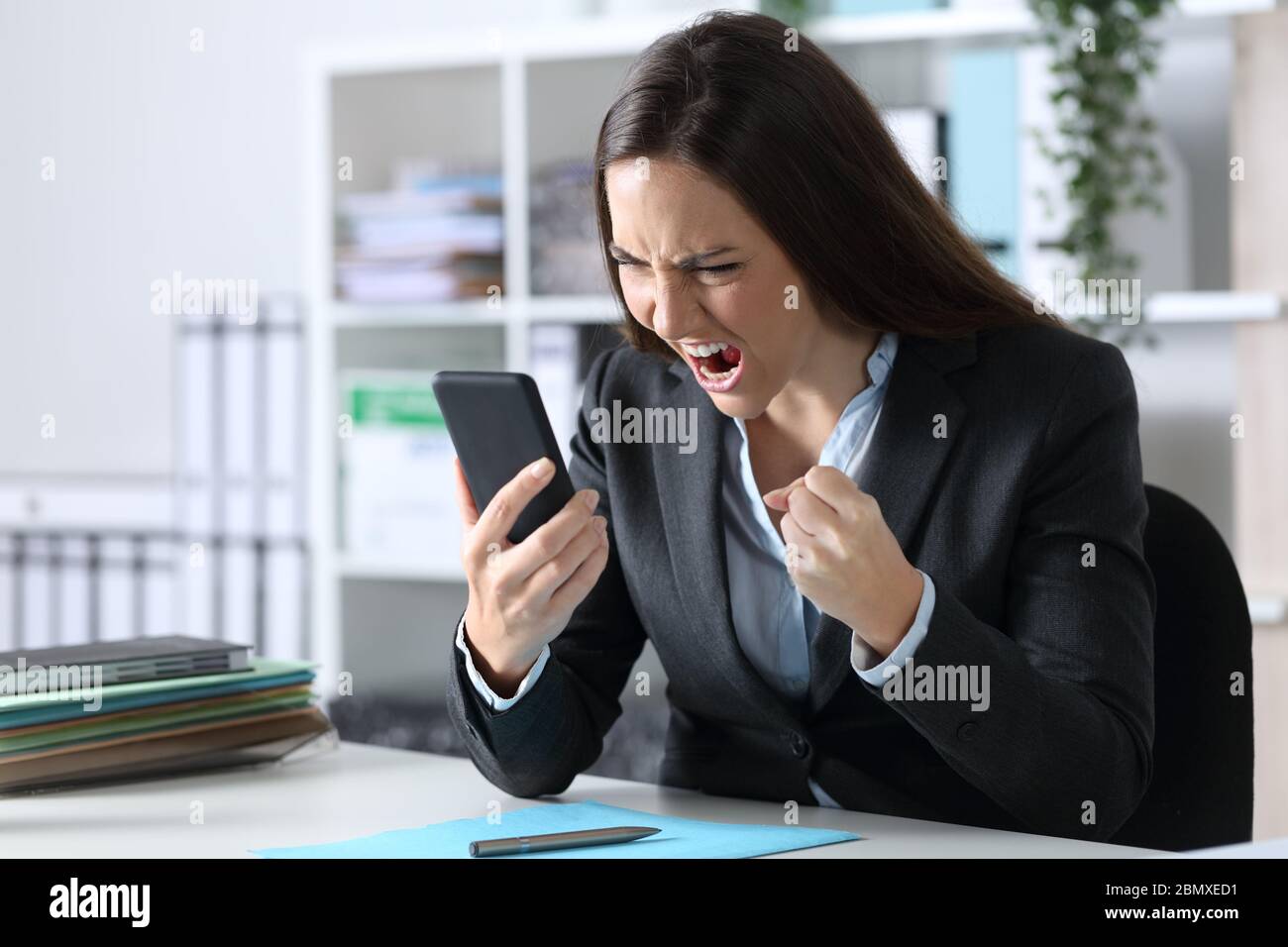Donna esecutiva arrabbiata che legge le cattive notizie sullo smartphone seduto su una scrivania in ufficio Foto Stock