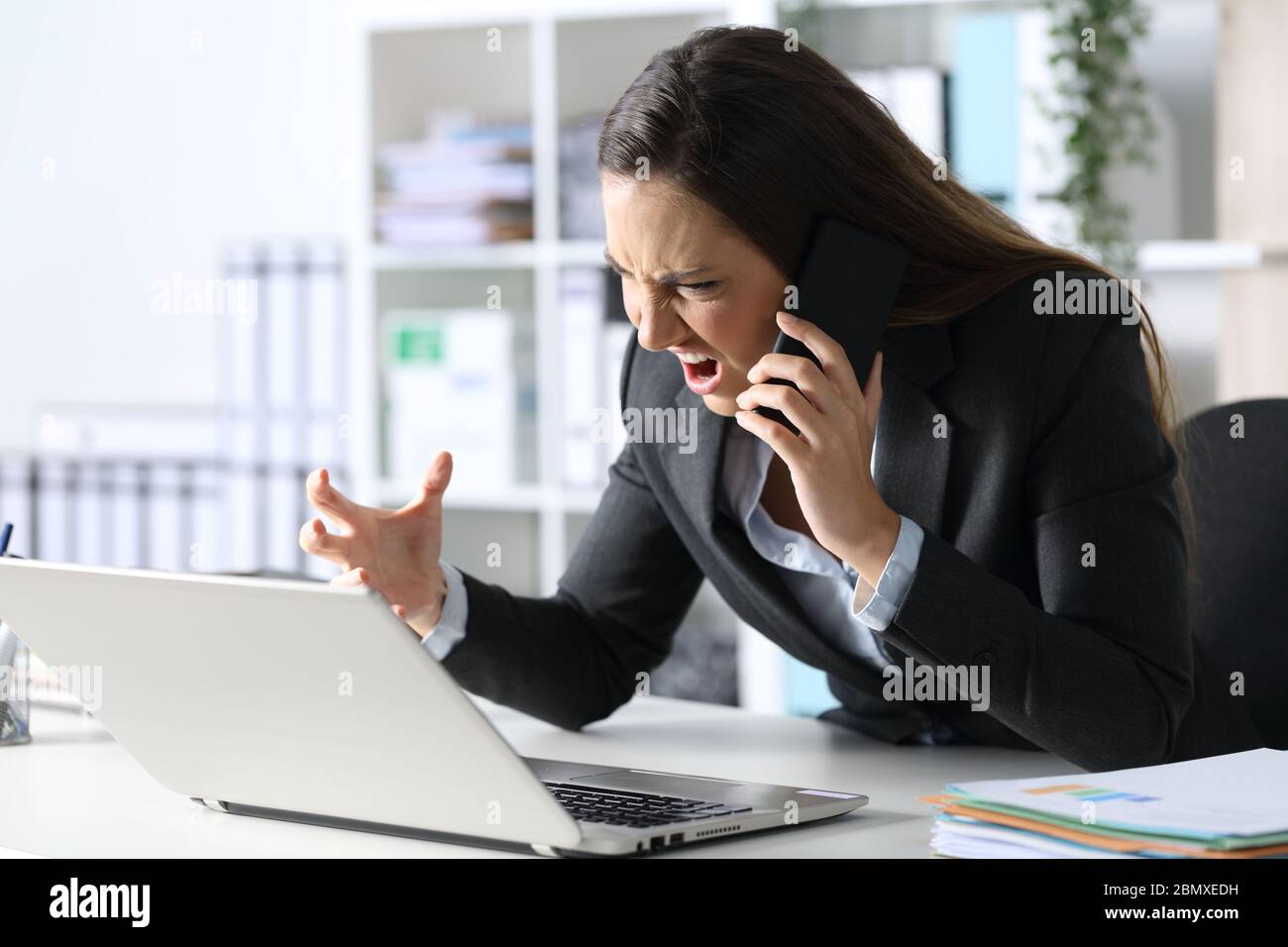 Donna executive arrabbiata che chiama sullo smartphone guardando il laptop seduto sulla sua scrivania in ufficio Foto Stock