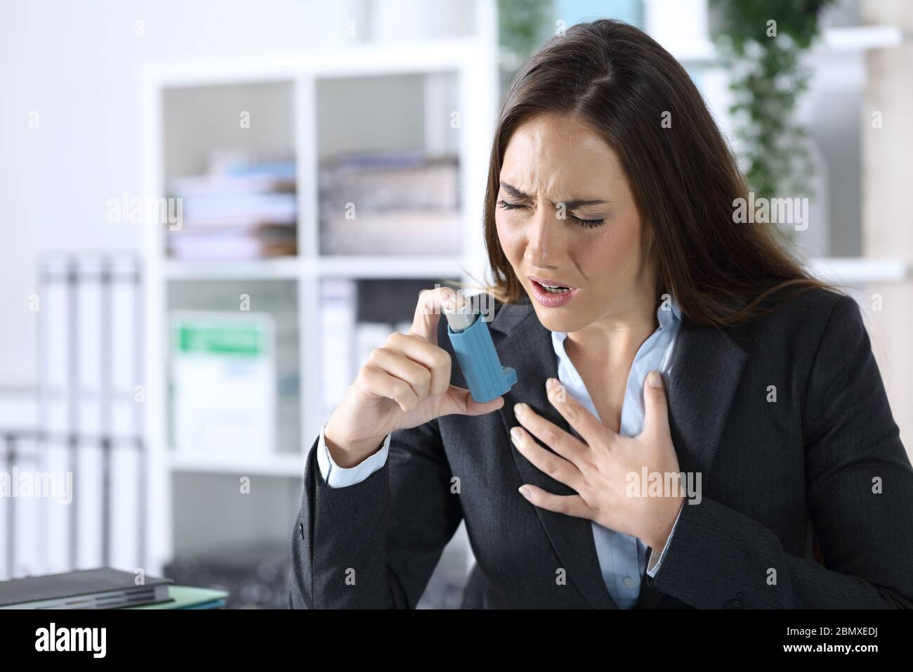 Donna executive che tiene inalatore con attacco di asma soffocante seduta su una scrivania in ufficio Foto Stock