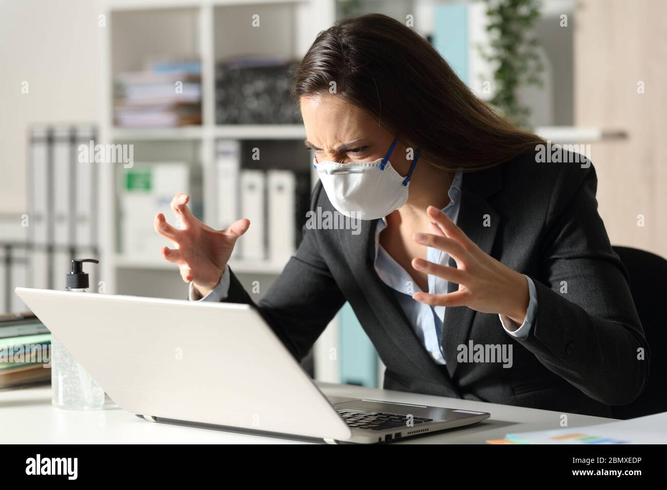 Donna executive arrabbiata che guarda il notebook evitando il covid-19 seduto sulla sua scrivania in ufficio di notte Foto Stock