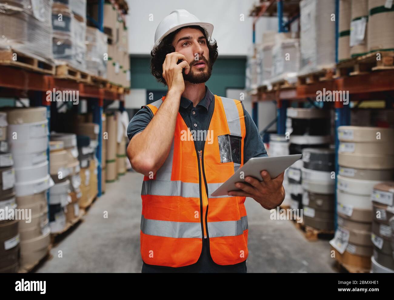 Tensed warehouse manager che tiene un tablet digitale mentre parla con un telefono cellulare che ha un aspetto stressato Foto Stock