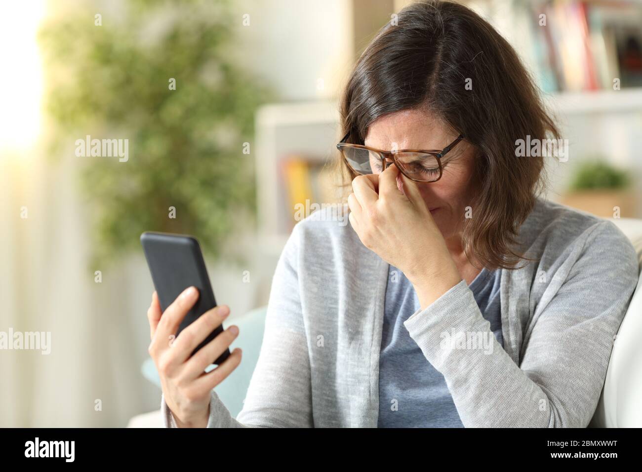 La donna adulta soffre di affaticamento degli occhi con occhiali e il telefono che tiene seduto su un divano a casa Foto Stock