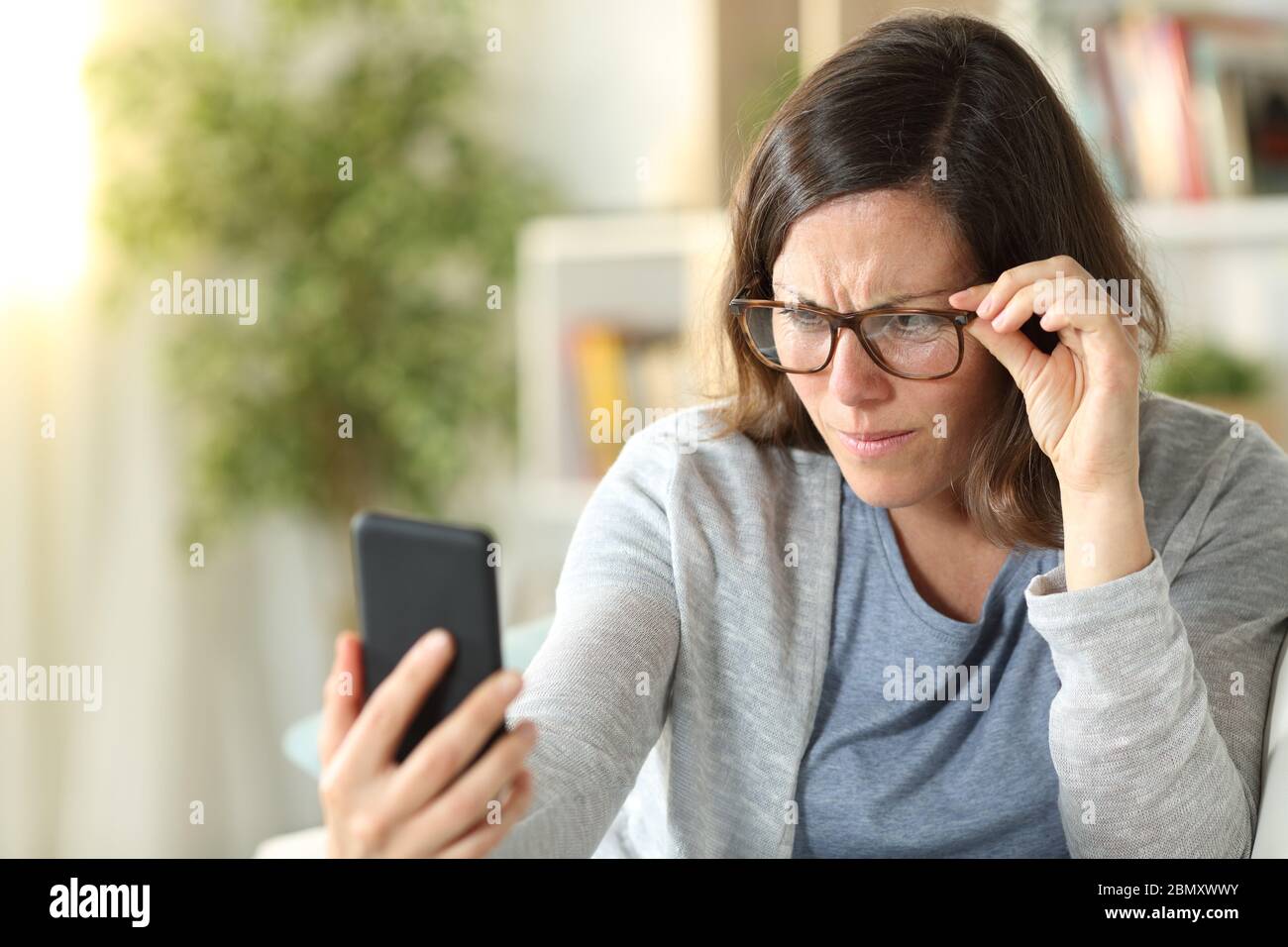 Donna adulta con problemi di vista indossare occhiali leggere su smartphone seduto su un divano a casa Foto Stock