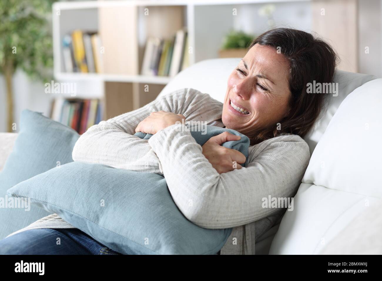 Triste donna adulta che piange seduta sul divano che tiene il cuscino nel soggiorno a casa Foto Stock