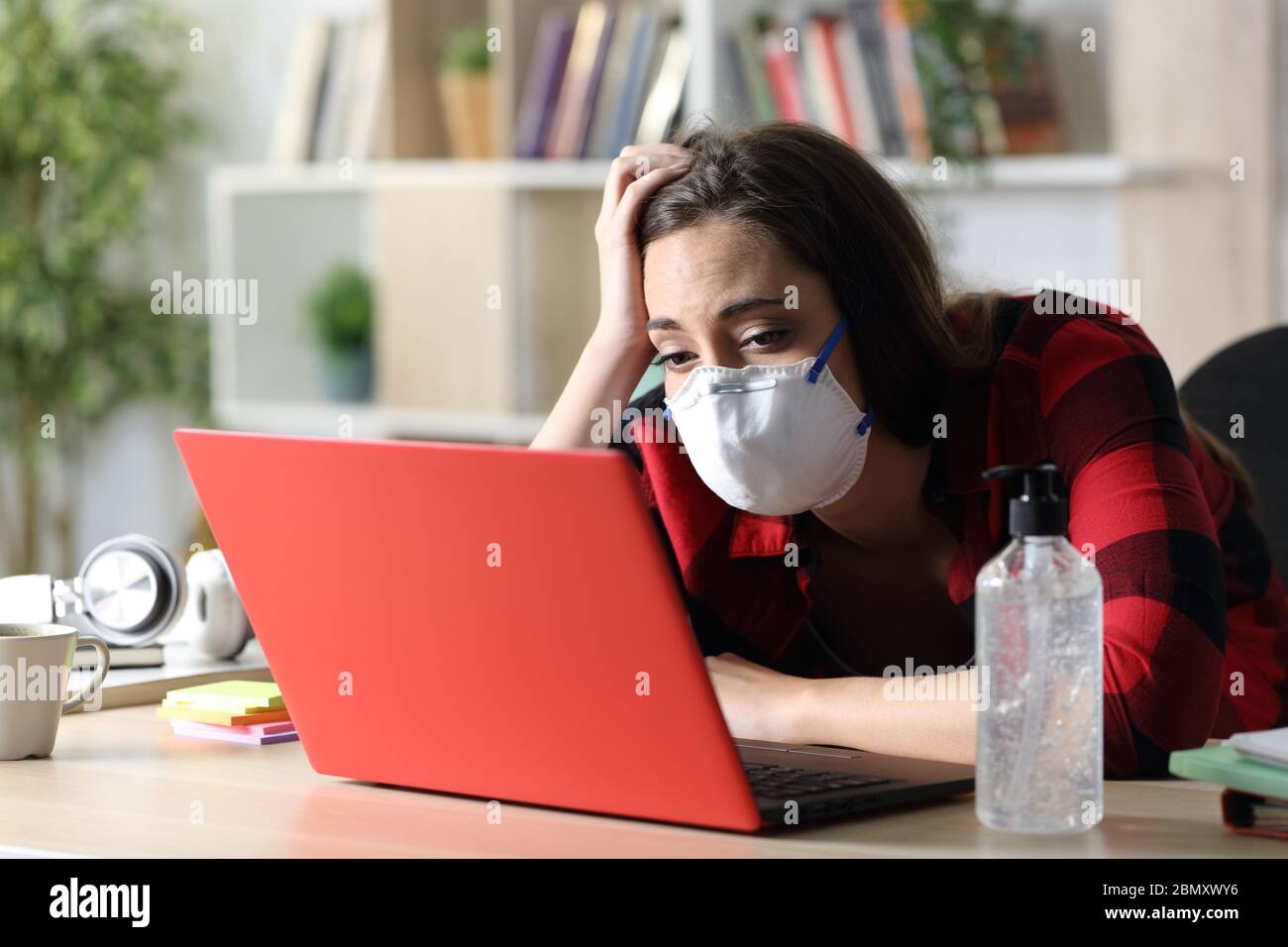 Studentessa annoiata con maschera che guarda il computer portatile seduto su una scrivania in confino al coronavirus a casa Foto Stock