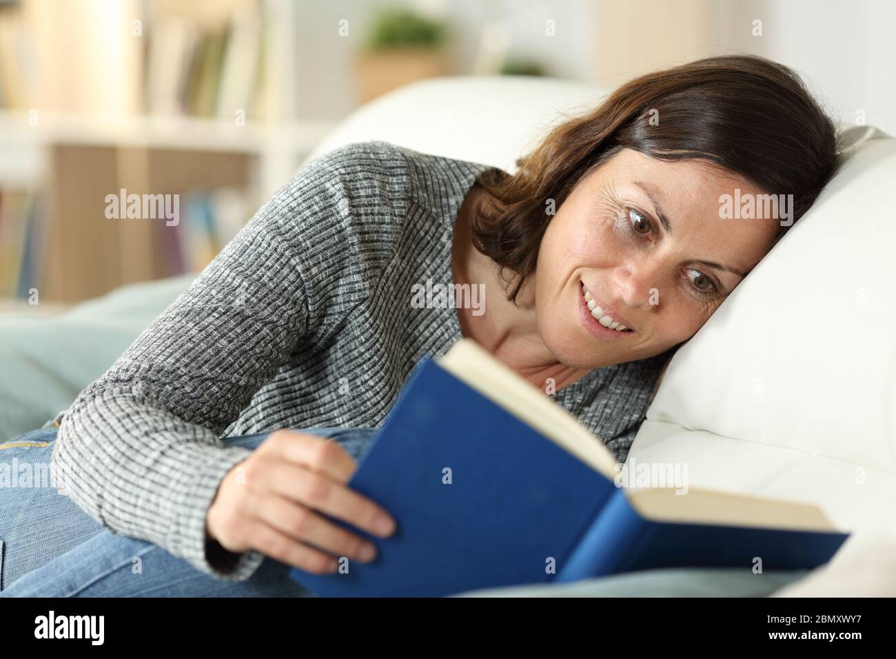 Felice donna di mezza età che legge libro di copertura duro seduta su un divano a casa Foto Stock