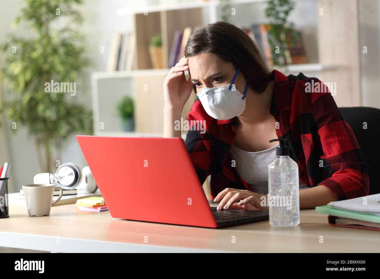 Triste studente con maschera protettiva che legge cattive notizie sul coronavirus su un computer portatile seduto su una scrivania a casa Foto Stock