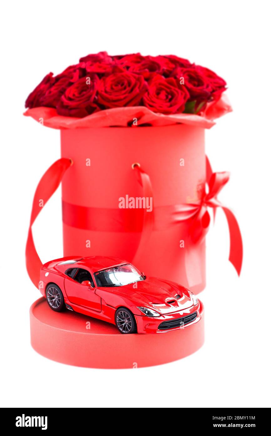 Giocattolo rosso moderno auto contro rose rosse di lusso in carta cappello scatola su sfondo bianco. Regalo romantico per donna Foto Stock