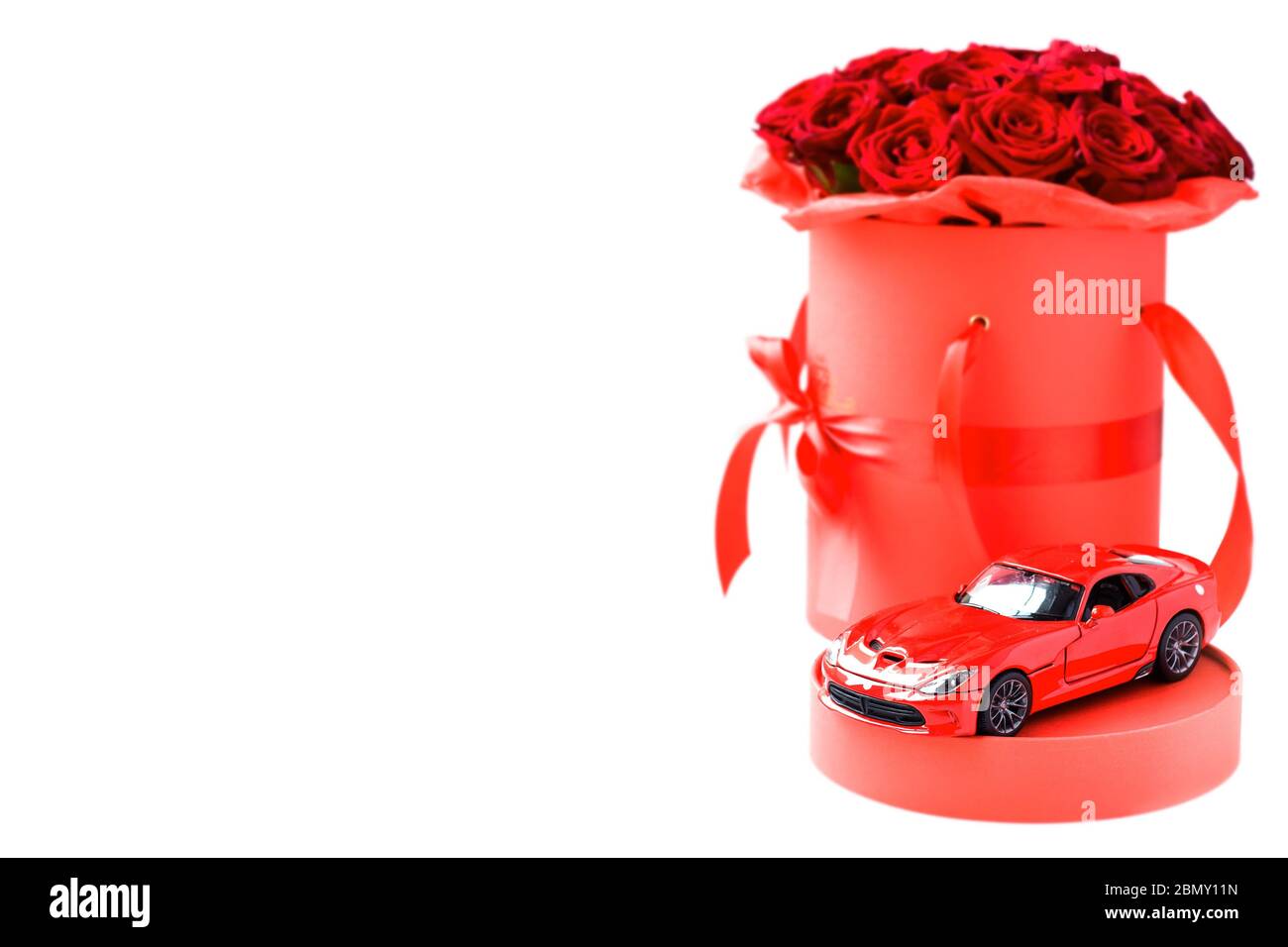 Giocattolo rosso moderno auto contro rose rosse di lusso in carta cappello scatola su sfondo bianco. Regalo romantico per donna Foto Stock
