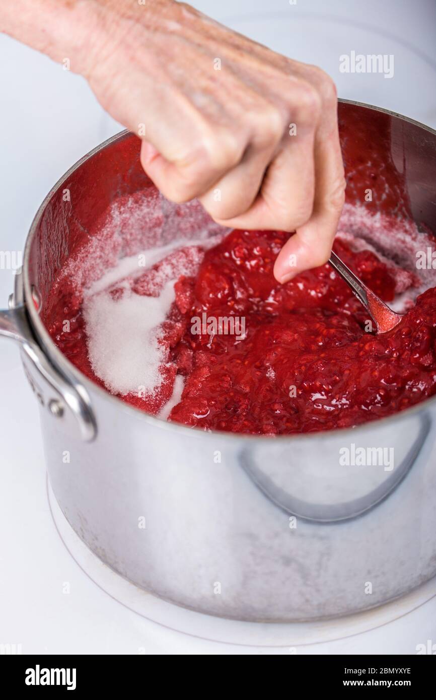 Donna che mescola lo zucchero in una pentola di lamponi schiacciati in marmellata-fare. Lo zucchero estrae l'acqua dalla frutta e forma legami chimici che "legano" il Foto Stock